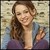 A static pixelated image of a person with long brown hair wearing a blue jacket over a purple top, holding a small object near their face.