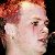 Pixelated close-up of a person's face with red hair, visible ear, and dark eye, set against a dark background.