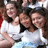 Three women in white tops pose together with one woman's arm around another, all smiling against a blurred background.