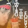 A person wearing a red bandana and dark hat holds a red and white object while standing before a background with large white text reading "SWING THE FOGS".