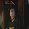 A person wearing a black cowboy hat and dark shirt with a colorful graphic on the chest, seated in a dimly lit room with dark curtains and a red chair.