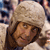 A pixelated close-up of a person wearing a light brown fur hat and dark brown clothing, with a blurred background of indistinct shapes and colors.