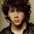 Pixelated portrait of a person with voluminous dark curly hair, wearing a dark jacket over a light shirt against a muted beige background.
