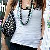 A person wearing a white tank top, dark pants, a long beaded necklace, and a black studded shoulder bag.