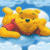 Pixelated yellow bear with brown nose and green eyes, wearing red shirt, lying on blue background with white clouds.