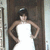 A person in a white dress with hands on hips stands against a dark background, wearing a small headpiece.