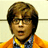 Pixelated static image of a person with brown hair wearing glasses, an orange jacket over a blue and white checkered shirt, and a yellow tie against a light background.