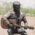 A pixelated figure in dark clothing holds a brown guitar-like instrument against a blurred green and beige background.
