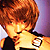 Pixelated static image of a person with curly brown hair wearing a dark top against a purple background, rendered in low-resolution 256x256 pixels with warm color tones and visible dithering.