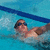 Pixelated image of a person swimming underwater with goggles, wearing dark swim cap and brown trunks against blue water background.