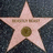 A light pink star-shaped icon with a dark brown circular center, set against a dark blue pixelated background.