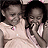 Two children in light-colored sleeveless dresses pose closely together against a dark background, one child's hand raised near the other's face.