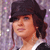 A person with light brown skin wears a dark flat cap and a white-collared shirt against a pixelated purple background with scattered light spots.
