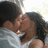 Two figures in close physical contact, one with dark hair and light skin wearing a white shirt, the other with dark hair and brown skin, against a light background.