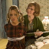 Two women stand side-by-side in a room with patterned wallpaper; the woman on the left wears a brown plaid dress with white trim and has curly blonde hair, while the woman on the right wears a green cardigan over a patterned top.