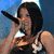 A person with short dark hair holds a microphone near their mouth, wearing a dark sleeveless top and light-colored skirt, with a bright green rectangle containing text in the lower right.