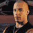 Pixelated static image of a bald man with light skin wearing a black shirt, set against dark and brown background elements.