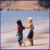 Two figures stand on a beach holding hands, one in dark clothing and the other shirtless with light hair against a sky-blue background.