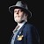 Pixelated static image of a person in profile wearing a gray hat and dark blue jacket with light-colored shirt, set against black background.