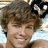 A pixelated close-up of a person with light brown curly hair, smiling slightly against a blurred background.