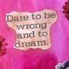 A static image shows a light brown, irregularly shaped text box with dark brown text reading "Dare to be wrong and to dream." on a pink background with faint floral patterns.