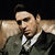 A man in a dark suit and white shirt sits on a light brown couch, his head and gaze shifting across three