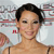 Pixelated close-up of a person with short dark hair, light skin, and a neutral expression, wearing a strapless top against a blurred background with red and gray tones.