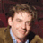 A pixelated image of a person with light brown hair wearing a brown jacket over a light blue shirt, set against a red background with pink text.