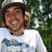 A person wearing a white helmet and white t-shirt with blue and yellow graphic, smiling against green foliage background.