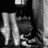Static grayscale image showing a stylized figure with long limbs and abstract shapes against dark background.