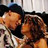 Two figures in close physical contact, one with light skin and dark hair wearing a white garment, the other with darker skin tones and brown hair against a blurred background.