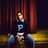 Pixelated image of a person in dark clothing with blue 'P' on chest, seated against red curtains and yellow bench, overlaid with bold black text on bright red background.