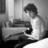 Black-and-white pixelated image of a person with short dark hair wearing a light sleeveless top, seated at a desk with a computer monitor and keyboard.