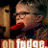A pixelated image of a person with short hair and glasses, mouth open in an exaggerated expression, red lips visible, wearing dark clothing against a dark background.