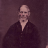 Pixelated image of a person with a bald head, light skin, and a dark V-neck garment against a dark purple background.