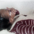 Pixelated image of a person in dark clothing with maroon accents, head tilted back, mouth open, and eyes closed against light background.