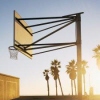A basketball hoop with a backboard and net stands against a clear sky, palm trees silhouetted in the background at sunset.