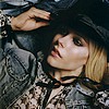 A person with short blonde hair wears a dark hat and patterned jacket, hand raised near the hat, against a blurred background.