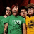 Four pixelated figures stand closely together, wearing green and yellow shirts with a white logo on the green shirt, against a dark background.