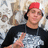 A person wearing a red cap and black shirt with silver graphic, making a peace sign against a patterned background.