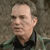 Pixelated close-up of a person with light skin tone wearing a dark collared shirt against a muted brown background.