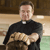 A pixelated image of a person in a black clerical shirt with a white collar, standing indoors with wooden and light-colored walls.