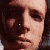 Pixelated close-up of a person's face with dark hair, visible eye, nose, and mouth, rendered in warm brown and beige tones with dark background.