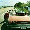 Rear view of a vintage convertible car driving on a highway with trees and open fields in the background.