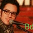 A person with dark hair and glasses sings into a microphone, wearing a patterned shirt against a red background with blurred lights.