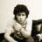 Pixelated static image of a person with dark curly hair wearing a light-colored shirt, seated at a desk with a computer monitor and keyboard.
