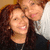 Two people with long curly brown hair pose closely together, one in a black top and the other in a white top, smiling at the camera.