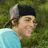 Pixelated image of a person in profile wearing a cap and light blue shirt against green foliage, with cyan text on solid green background.