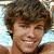 Pixelated close-up of a person with curly brown hair, light skin, and a smiling expression against a light blue background.