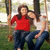 Two people sit on a swing in an outdoor setting, one wearing red top and dark pants, the other in white top and blue jeans.